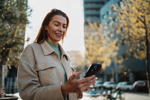Happy lady looking at smartphone screen holding cup of coffee outdoors. Modern woman using mobile internet for social network communication or shopping online. Authentic professional lifestyle.