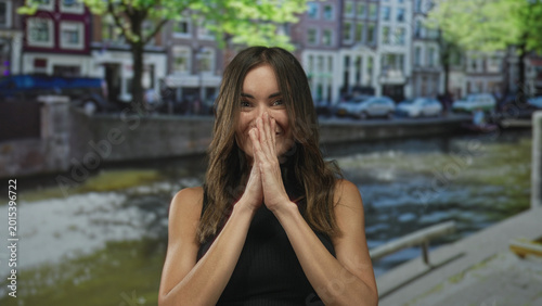 Fotografija Hispanic woman grins and raises hands in shrug beside sunlit water canal by historic building; excitement