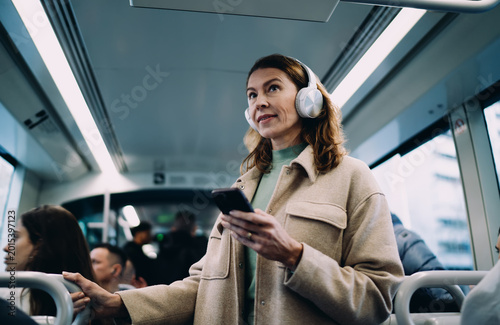 Schilderij op canvas Caucasian middle aged woman holding handle in tram, using smartphone, wearing headphones, looking forward, public transport commuter scene, modern digital lifestyle, authentic urban life