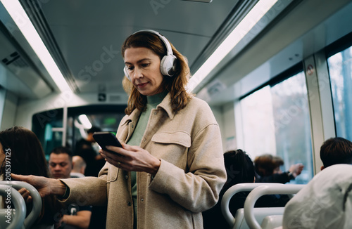 Bild auf Leinwand Caucasian middle aged woman standing inside tram, using smartphone app, wearing white headphones, commuting in public transport, holding handle, digital lifestyle, looking at phone screen