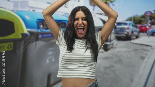 Foto Woman with arms raised above head beside large recycling bin, mouth open and tongue out in a spirited pose on a crowded urban street; playful joy