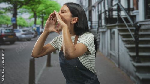 Φωτογραφία Woman in denim apron cupping hands to mouth shouting on a city street by stoop and parked cars with bollards visible; urgent call