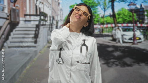Tableau sur toile Woman doctor in white coat with stethoscope clutching her neck and grimacing, wearing yellow glasses on a leafy street beside brownstone steps and parked car; stress