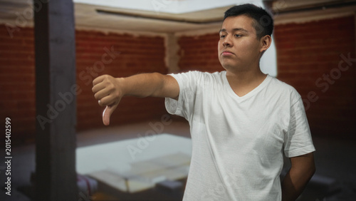 Fotografi Man showing thumbs down with outstretched arm and visible thumb inside building interior with brick wall; disapproval critique