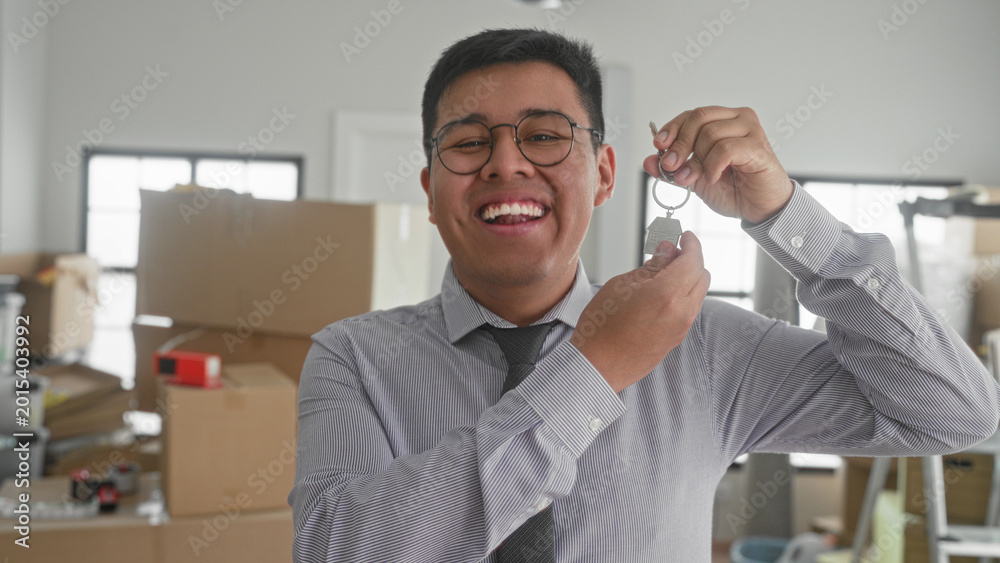 Fototapeta premium Man holding a house key and smiling in a building surrounded by moving boxes and ladder; homeownership joy.