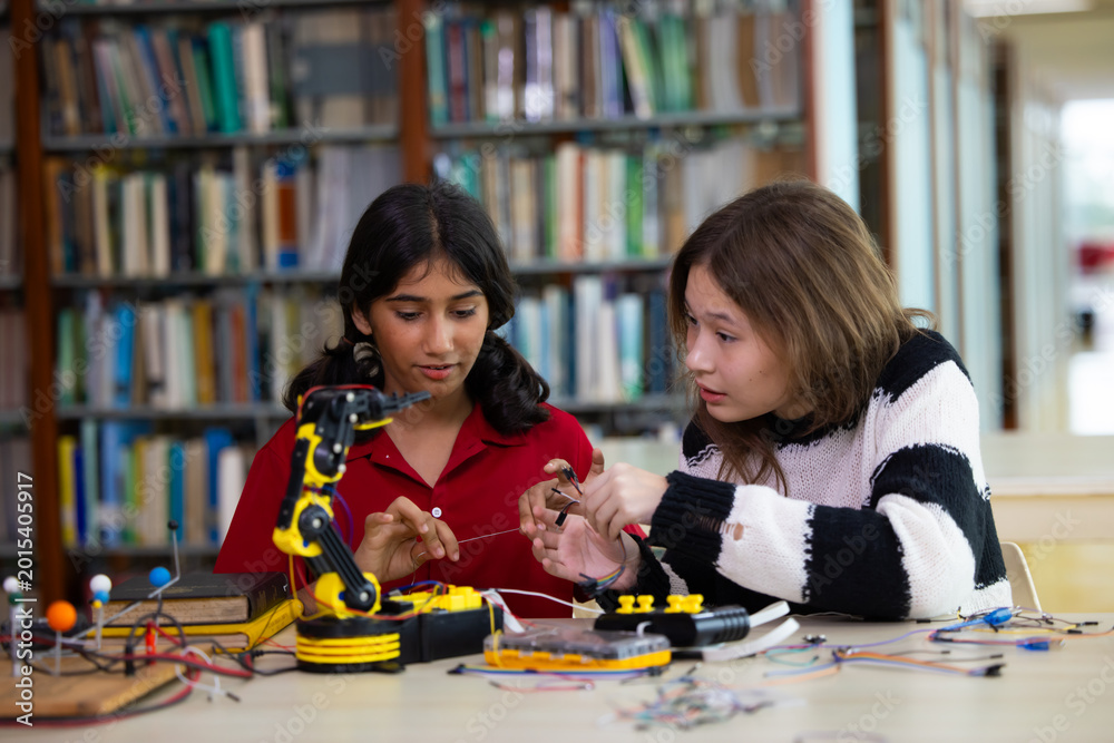 Fototapeta premium Indian girls focus intently on robotic arm project. hands adjusting wires and parts. The library setting suggests learning or innovation. Their expressions show curiosity. Education, Back to school