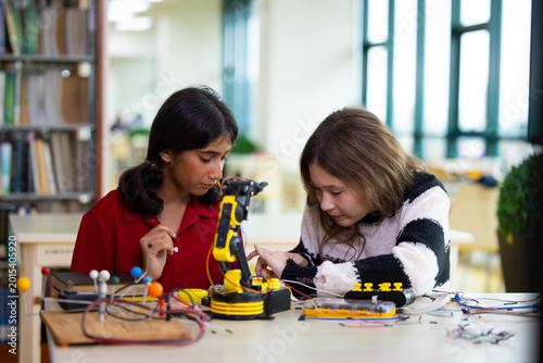 Indian girls focus intently on robotic arm project. hands adjusting wires and parts. The library setting suggests learning or innovation. Their expressions show curiosity. Education, Back to school