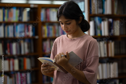 Young Indian teenage girl smiles and selects blue book. Her fingers gently touch the cover's edge. Behind her, wooden shelves hold many books. Education, Back to school