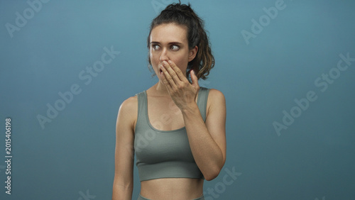 Foto Young hispanic woman wearing a gray tank top and bare midriff covers mouth with hand in blue studio setting; surprise