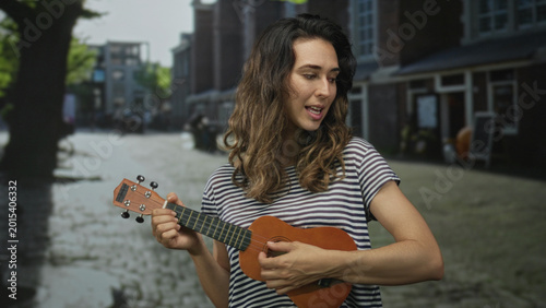 Fotografia Woman strumming a small wooden ukulele on a sunlit cobblestone street wearing striped shirt and smiling at instrument; music joy