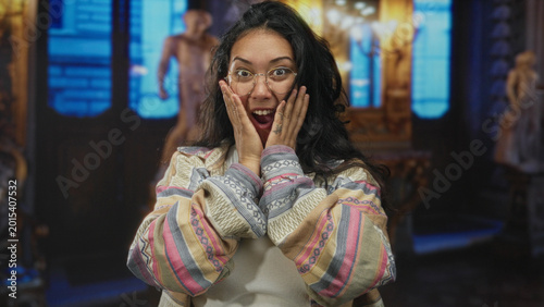 Bilde på lerret Woman hands on head laughing with mouth open in museum building, wearing glasses, hoop earrings and patterned shirt; joy discovery