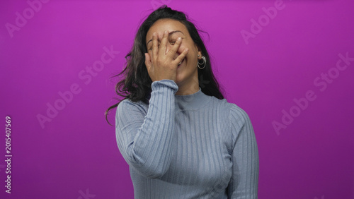 Photos Woman young laughing with hand covering face and open mouth in purple studio wearing blue sweater; joy confidence