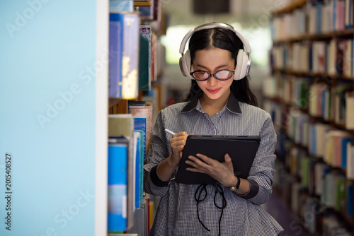 Young Asian woman smiles while listening to music in library. She wears glasses and headphones, Bookshelves line both sides of the aisle behind her. Soft light illuminates her focused. Education
