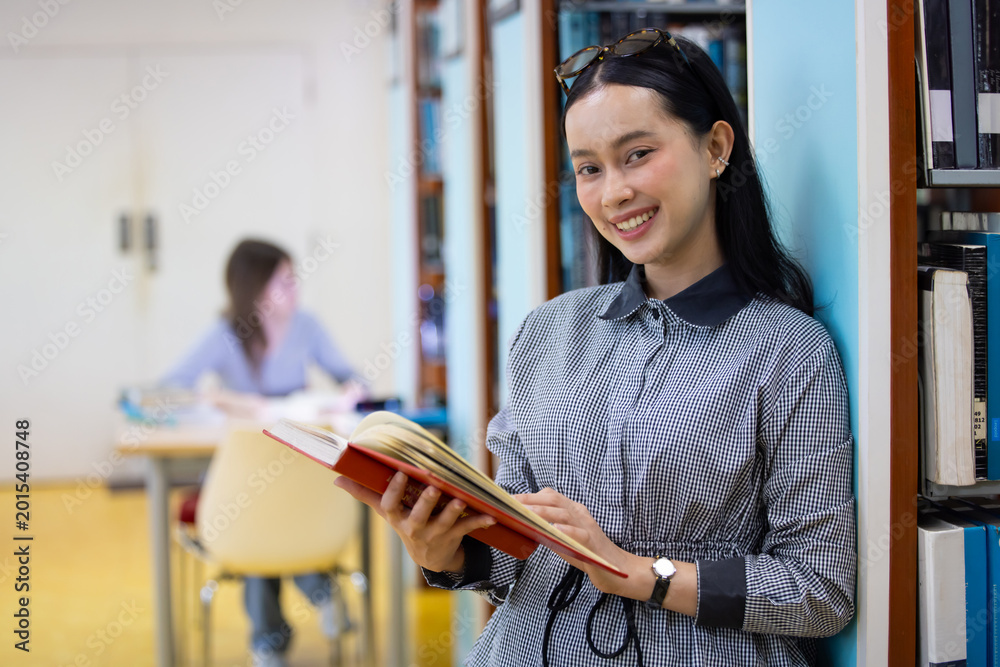 Fototapeta premium Young Asian woman studies in quiet library. Books line tall shelves behind. she holds book, eyes focused. Serene atmosphere, calm concentration. Ideal for educational or study-themed projects