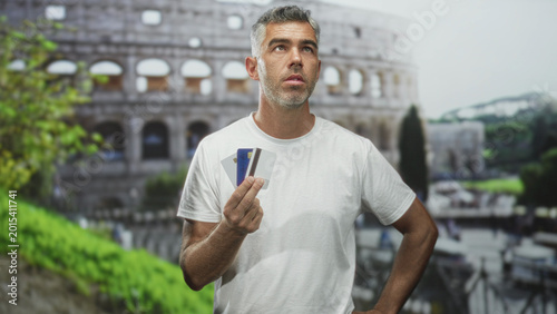 Photography Man holding creditcard and debitcard in hand at historic building colosseum, facing camera with pensive look; financial anxiety