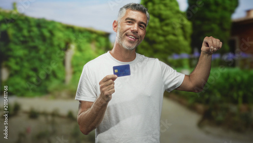 Quadro su tela Man holding a creditcard with his hand, showing the card while standing in front of a building, looking uncertain and reflective; financial concern