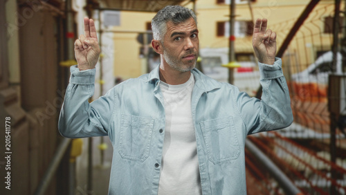 Fotografie Man raises two fingers on each hand in a street beside scaffolding and a building facade, wearing a denim jacket and white tshirt; playful confidence