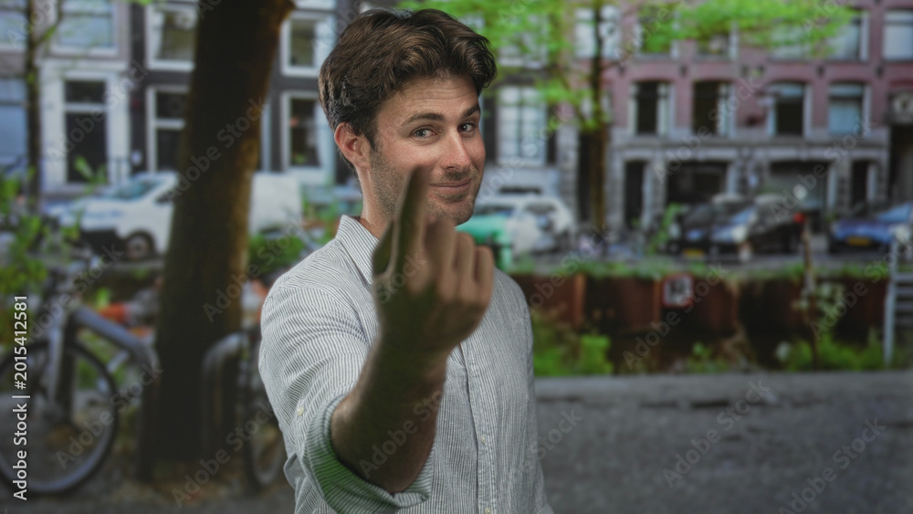 Fototapeta premium Young hispanic man shows middle finger to camera on a city street with bicycles and canal; defiance attitude.
