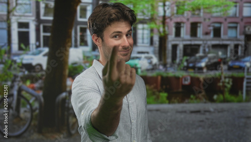 Fotografie Young hispanic man shows middle finger to camera on a city street with bicycles and canal; defiance attitude