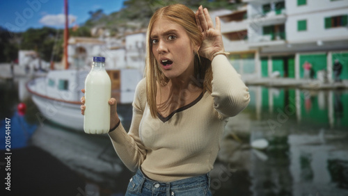 Obraz na plátne Young redhead woman holding a milk bottle cups hand to ear by boats and quay in a marina harbor with waterfront buildings; curiosity discovery
