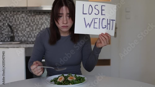 Young woman forces herself to eat healthy food while holding lose weight sign showing pressure of diet culture body standards and unhealthy obsession with thinness