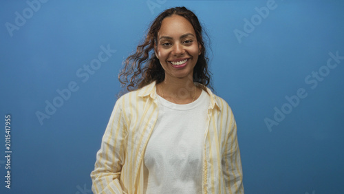 Foto Woman with hand covering eyes and hand over mouth wearing white t shirt and yellow striped shirt in studio blue backdrop, mid gesture portrait; playful candid