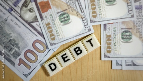 Foto "DEBT" are arranged diagonally over a collection of one-hundred-dollar bills, representing financial liabilities, borrowing costs, and economic obligations