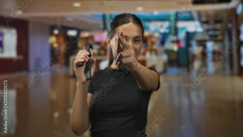 Canvas Print Young woman points finger like gun while holding a stun device at a mall kiosk, brunette in black shirt indoors center; self defense confidence