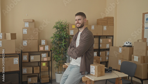 Quadro su tela Man with arms crossed leaning on a parcel at a packing table surrounded by stacked cardboard boxes and shelving in a building; small business confidence