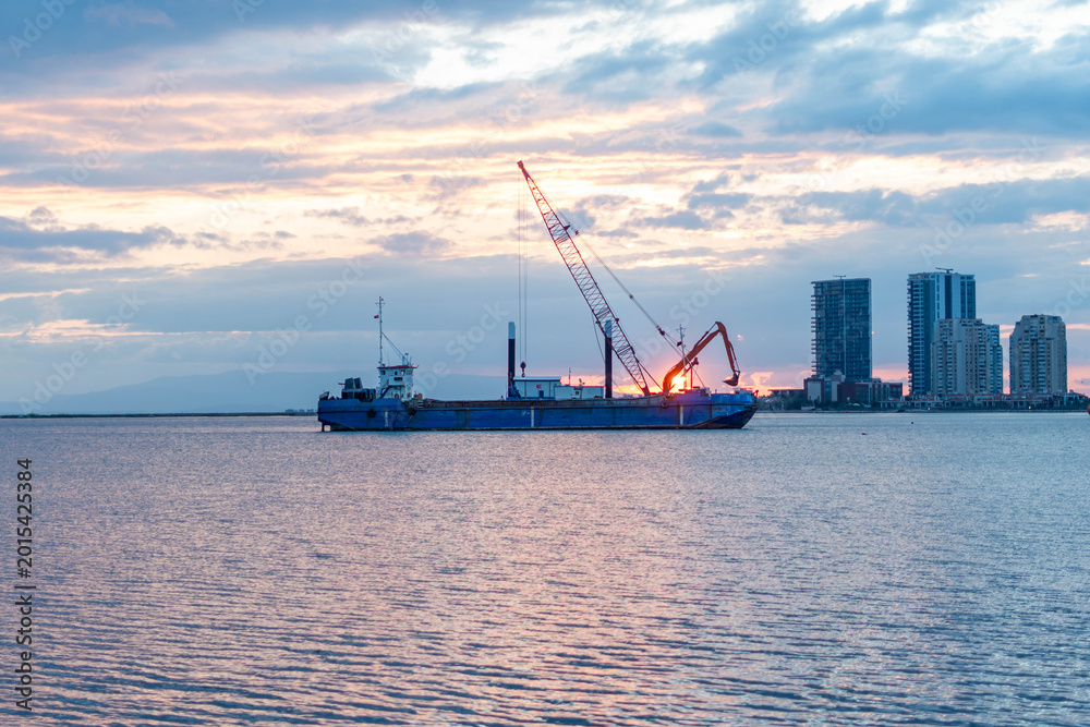 Fototapeta premium Construction barge with crane operates at sunset near modern city skyline