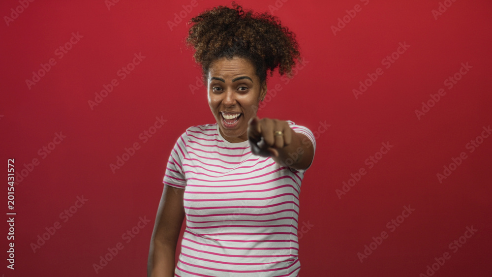 Αυτοκόλλητο Young african american woman laughing, pointing finger and covering mouth in red studio; playful teasing amusement