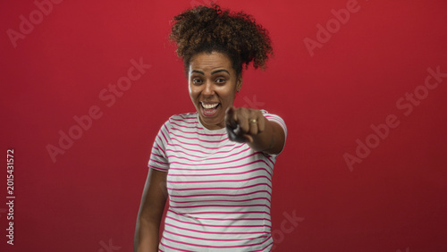 Quadro em tela Young african american woman laughing, pointing finger and covering mouth in red studio; playful teasing amusement