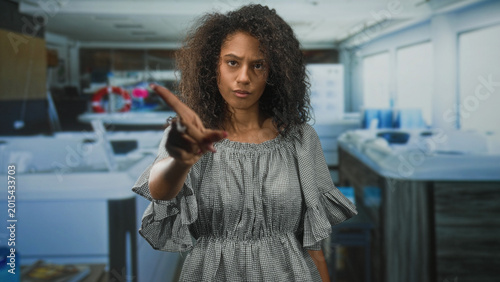 Obraz na plátně Teenage woman with curly hair in a gingham dress points index finger forward and briefly touches her nose in studio; determination