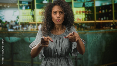 Photography Teenage woman with curly hair points finger toward camera amid bar counter inside building; anger confrontation