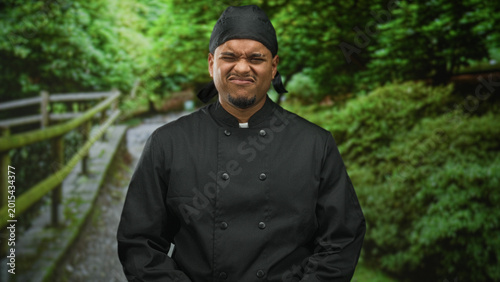 Obraz na plátně Chef man in black uniform, nose scrunched in a grimace on a forest path beside a wooden railing; disgust reaction
