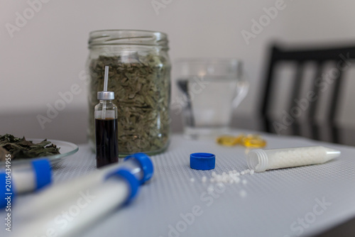 Fotografie Close-up of homeopathic remedies on table: small glass vial with herbal tincture, open tube with homeopathic pellets, jar of dried herbs and glass of water