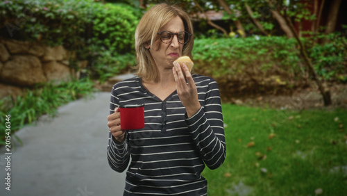 Fotografía Woman blonde in striped shirt holding red mug and muffin wrinkles nose and grimaces while inspecting muffin on street path; disgust morning routine
