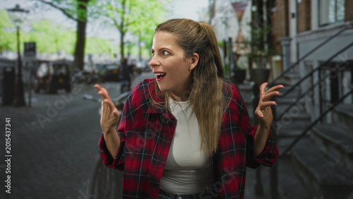 Φωτογραφία Young woman in red plaid shirt with hands raised and wide eyes, standing by a bicycle on a tree lined street; surprise explanation