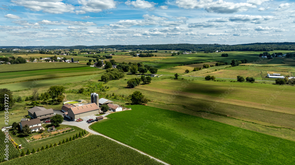 Naklejka premium A wide view of a rural area with green fields, farm buildings, and a clear sky. The setting is peaceful with a mix of crops and open land under clouds in the sky.