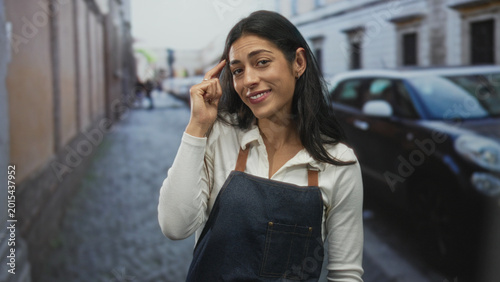 Fotografija Woman points finger to temple on a cobblestone street wearing denim apron and white shirt while standing near parked car; confidence cleverness wit