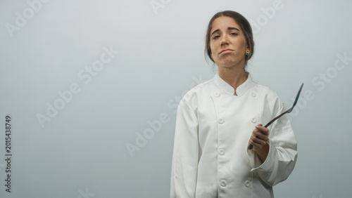Foto Young woman chef in white coat holds spatula with hand to forehead and open palm gesture in studio; frustration