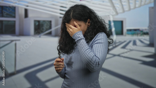 Photos Woman young brunette in blue turtleneck holding vape device with hand covering face in building with canopy and windows; anxiety personal struggle