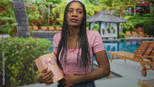 Fotografija Woman holds pink gift box with hand on abdomen and eyes rolled near a pool building; annoyance waiting uncertainty