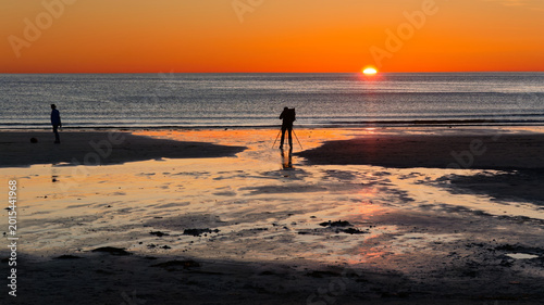Silhouetted figures on the beach: a photographer captures the sunset while a hiking companion waits. Lofoten Islands, Northern Norway. 