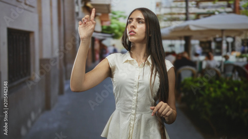 Fotografía Teenage girl in white dress pointing index finger with raised arm on a street cafe alley, long hair falling over shoulder; confidence