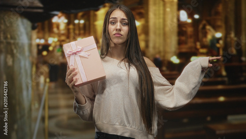 Foto Teenage woman holding pink gift box with ribbon, shrugging with one arm extended and pursed lips in an ornate building; ambivalence