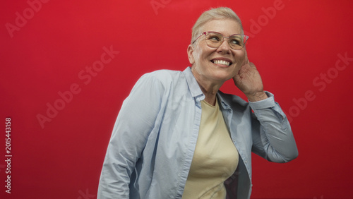Photos Woman cupping ear and leaning forward in a red studio wearing glasses and shirt, hand to ear gesture as if trying to hear; surprise attention