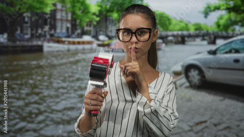 Tableau sur toile Young woman holding tape dispenser and finger to lips for silence on amsterdam street; quiet secrecy
