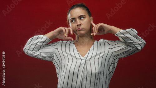 Valokuva Woman plugging ears with fingers in studio against a deep red backdrop, wearing a white striped blouse and looking upward with hands raised; frustration