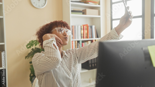 Bild auf Leinwand Woman holding smartphone smiling and raising arm to take selfie at desk with computer and bookshelves in building; workday joy